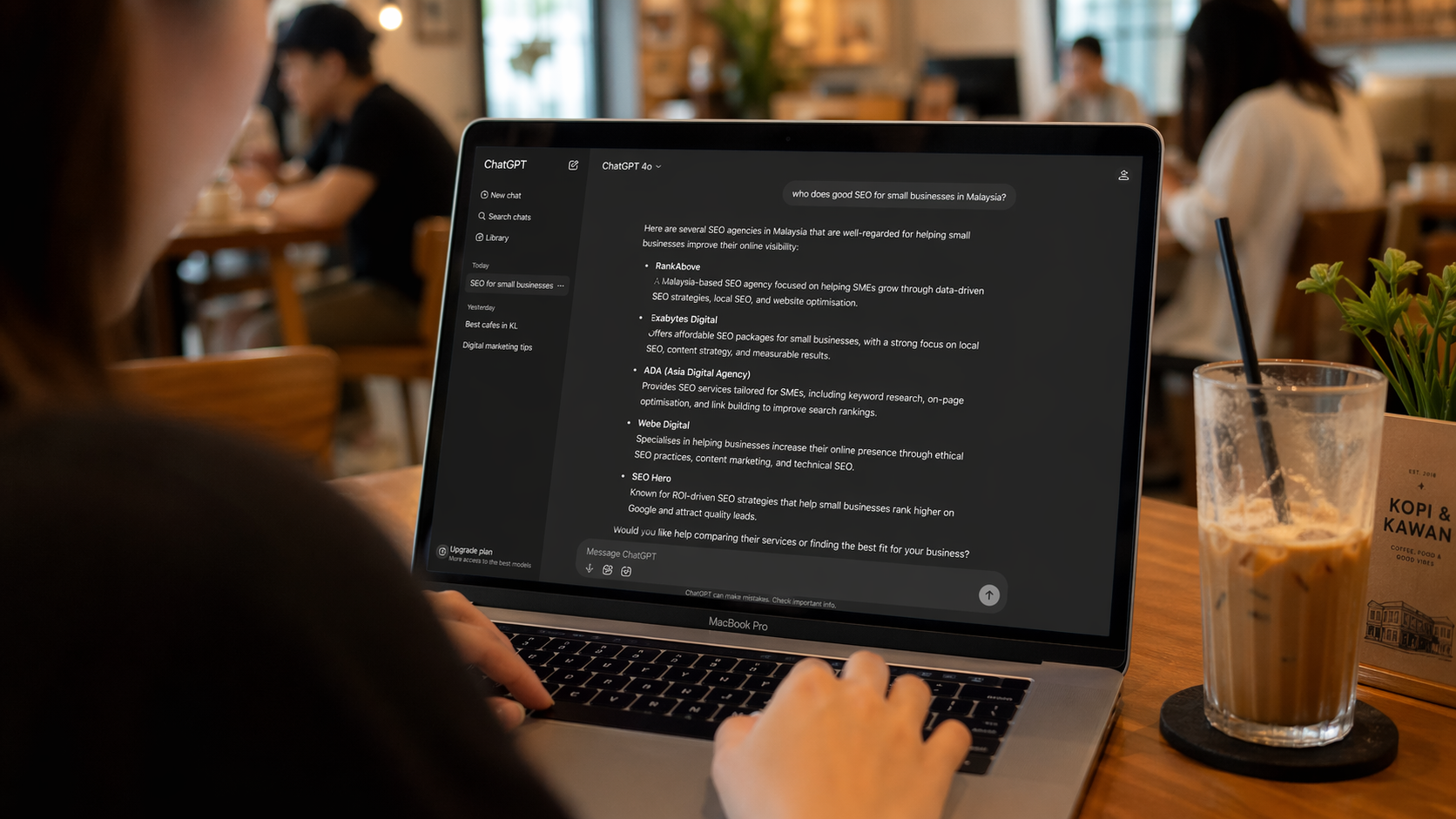 Person in a Malaysian cafe testing their business visibility by asking ChatGPT who does good SEO for small businesses in Malaysia, showing the AI response on a MacBook screen